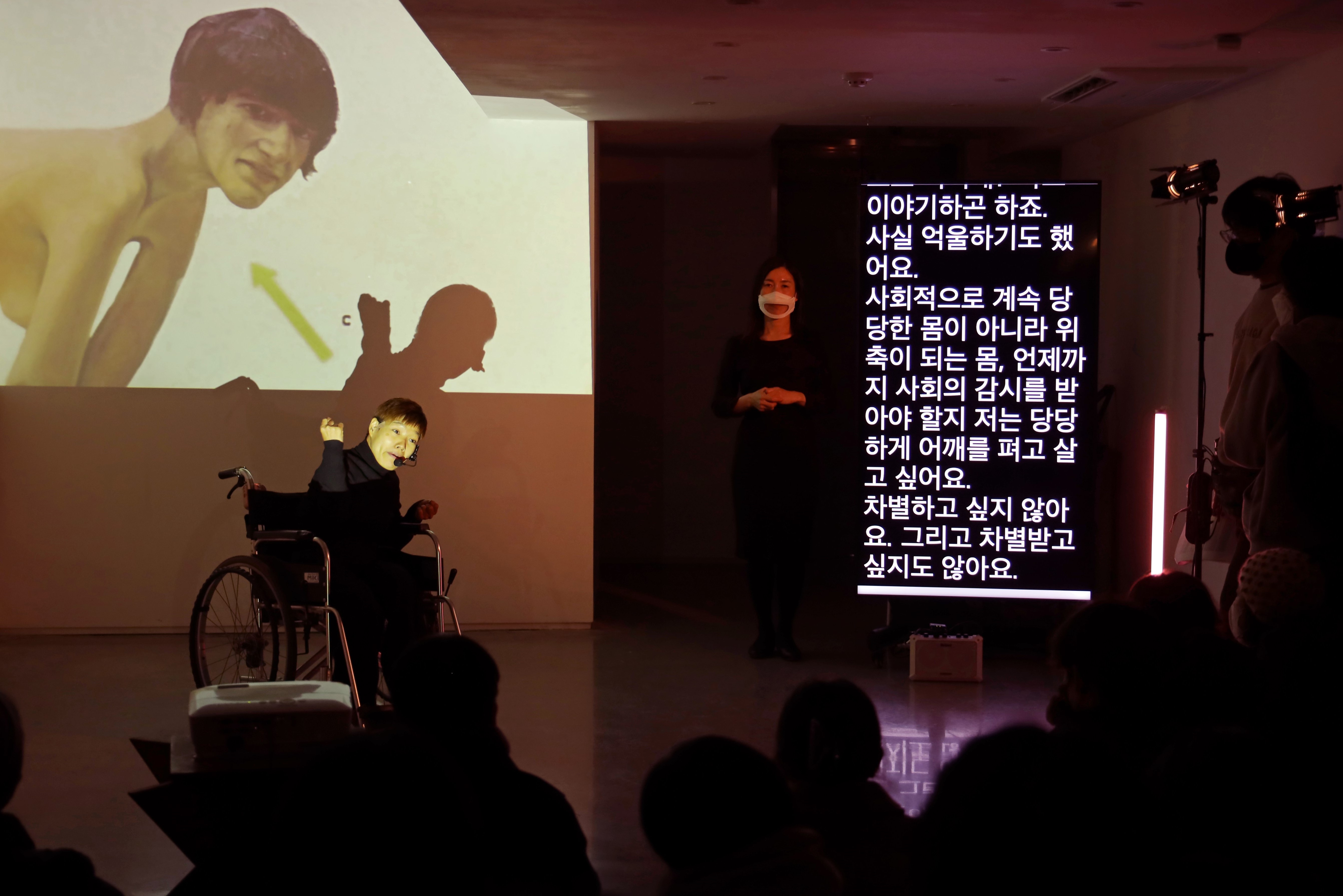 벽면을 가득 채운 빔 프로젝션 영상을 배경으로 휠체어를 탄 배우가 퍼포먼스를 하고 있다. 영상에는 한 사람의 몸 그림이 있다. 오른편에는 수어통역사와 자막이 있다. 자막에는 다음의 내용이 보인다. “이야기하곤 하죠. 사실 억울하기도 했어요. 사회적으로 계속 당당한 몸이 아니라 위축되는 몸, 언제까지 사회의 감시를 받아야 할지 저는 당당하게 어깨를 펴고 살고 싶어요. 차별하고 싶지 않아요. 그리고 차별받고 싶지도 않아요.”