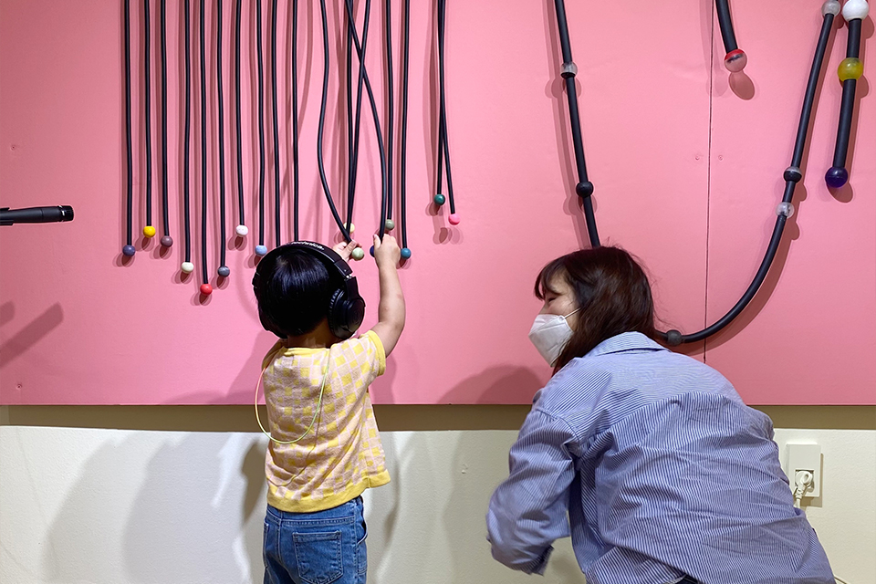 분홍색 벽면에 색색의 나무 공이 달린 검정 줄이 여러개 매달려 있다. 엄마와 아이가 헤드폰을 쓰고 이 작품을 만지며 체험하고 있다.