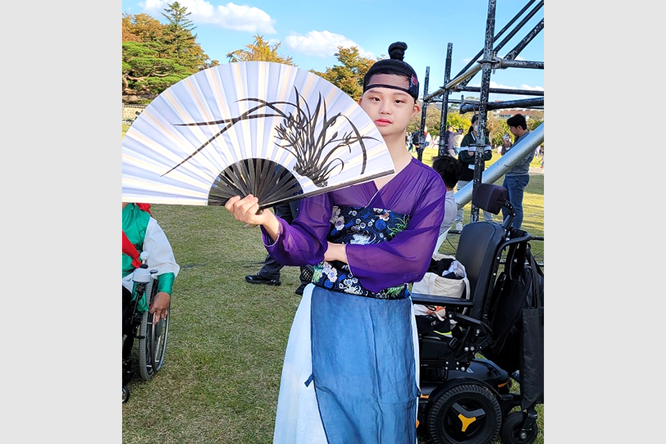 야외 축제 현장, 공연 분장을 한 장애 무용수. 보라색빛 얇은 저고리에 파랑색 계열 여러 겹의 하의를 입고, 허리에는 화려한 무늬가 그려진 두툼한 너비의 띠를 둘렀다. 머리에는 상투를 틀고 손에 커다란 합죽선을 들고 있다.