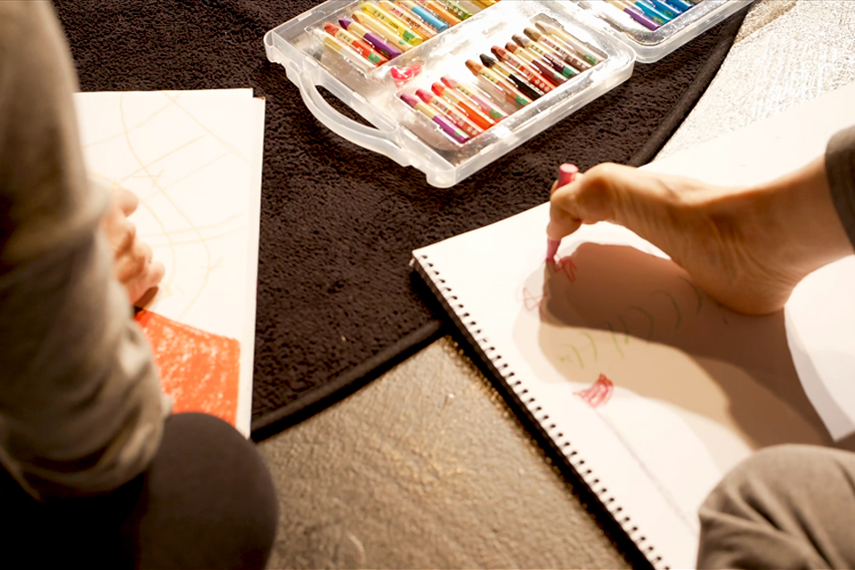 바닥에 크레파스가 놓여 있고, 각자의 스케치북에 그림을 그리는 두 사람. 한 사람은 발가락으로 붉은색 크레파스를 잡고 그린다.