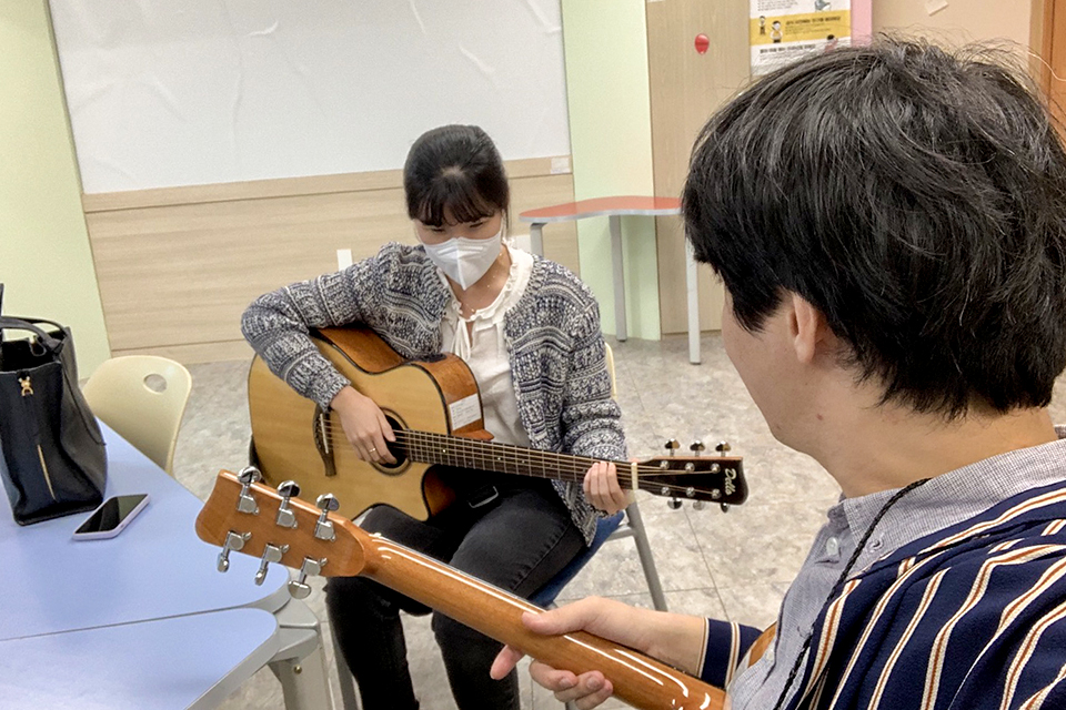시각장애인 참여자가 기타를 들고 앉아 있고, 그 앞에 기타를 든 강사가 이 모습을 지켜본다.
