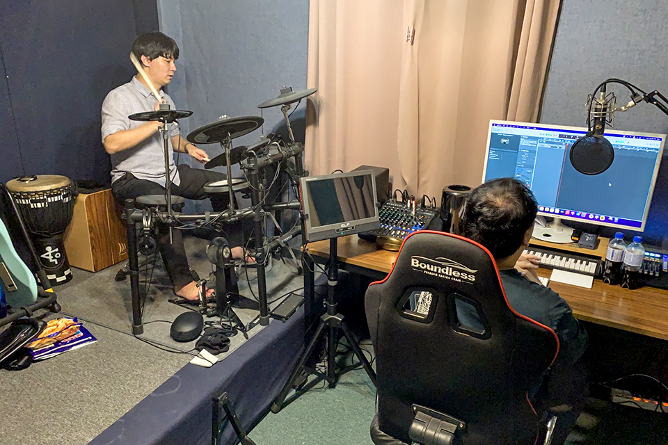 음악 연주와 녹음이 가능한 방 안, 한 명은 드럼을 연주 중이고 한 명은 컴퓨터가 놓인 테이블에 앉아 있다.