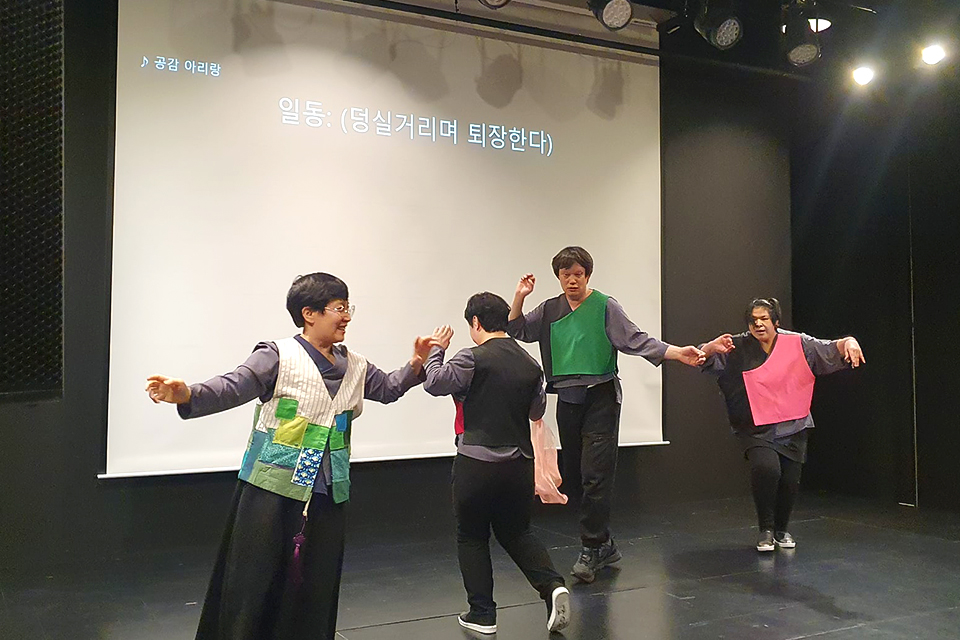 무대에서 네 명의 배우가 덩실거리며 춤추고 있다. 무대 벽면 자막에는 “공감 아리랑. 일동 : (덩실거리며 퇴장한다)라고 쓰여 있다.