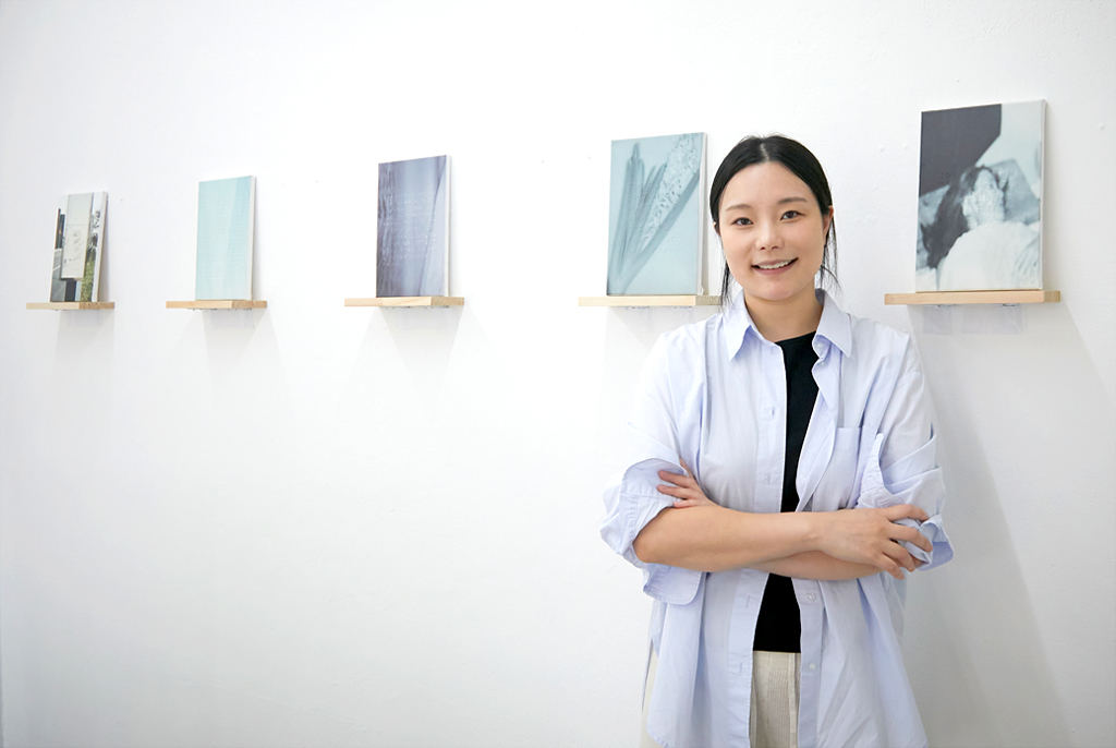 유다영 작가가 정면을 바라보고 팔짱을 낀 채 포즈를 취하고 있다. 뒤로 벽면에는 일렬로 사진작품이 걸려 있다.