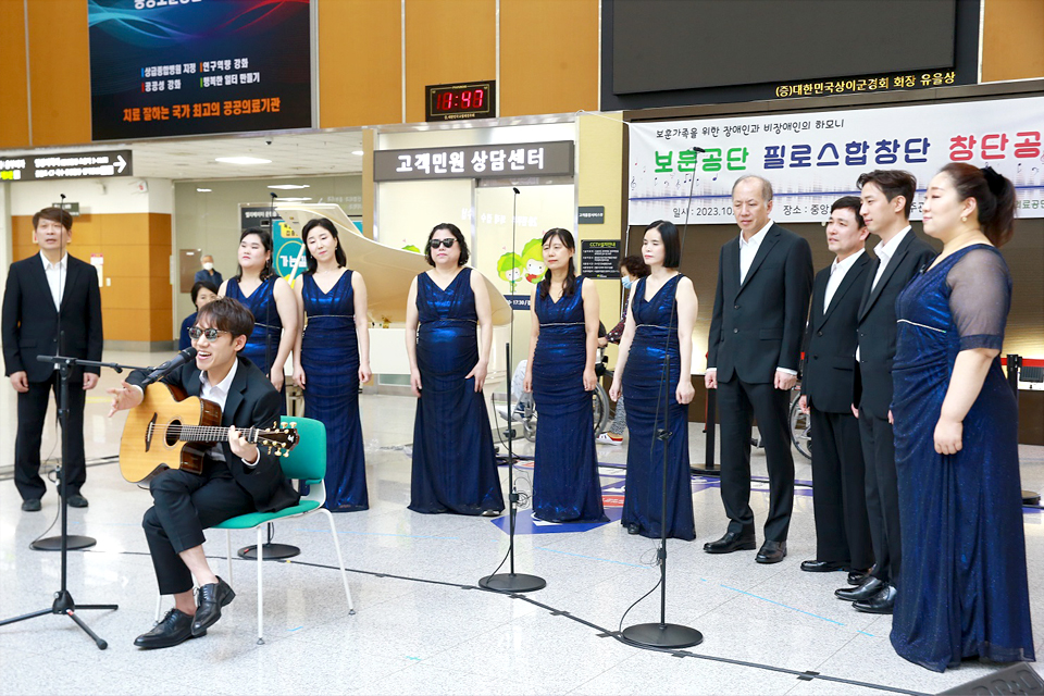 10여 명의 남녀 혼성합창단원이 빙 둘러 서 있고, 앞쪽 중앙에 신재혁 단원이 의자에 앉아 기타를 치며 노래하고 있다.