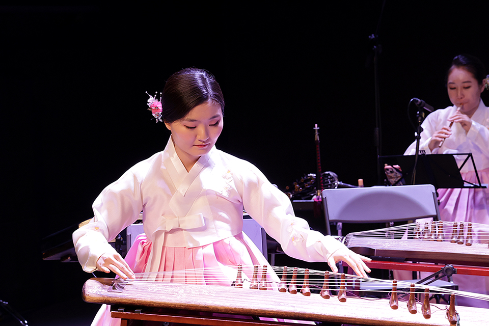 연분홍 저고리와 치마의 한복을 입은 김보경 연주자가 가야금을 연주하고 있다. 뒤편에 같은 한복을 입은 연주자가 피리를 연주하고 있다.