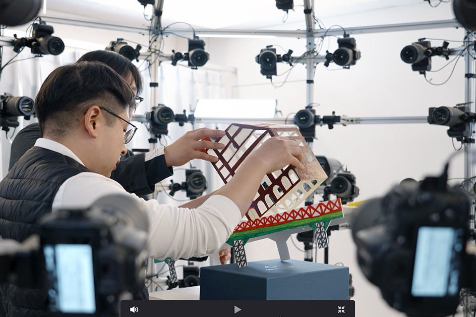 구조물에 수많은 카메라가 매달려 있고, 그 안에서 두 사람이 조형물을 손에 들어 3D 스캔하고 있다.
