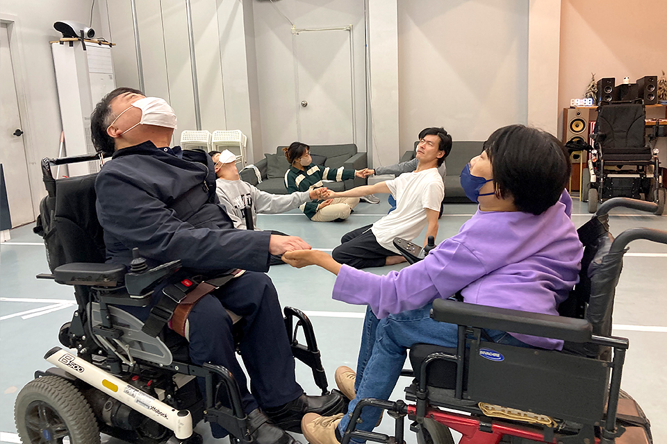 배우들이 두 명씩 짝을 지어 한 손을 마주 잡고 몸을 뒤로 젖히며 지탱하고 있다. 맨 앞 한 쌍은 휠체어를 타고 있다.