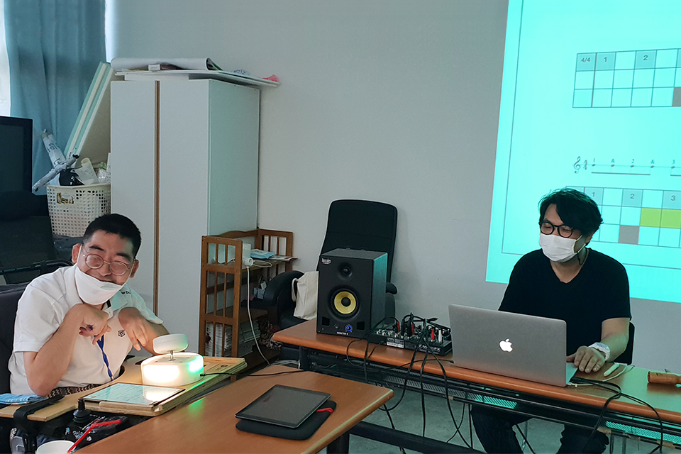 천필재 대표와 다른 한 사람이 책상 앞에 앉아 있다. 천필재 대표 앞에는 노트북 등 장비가 놓여 있고, 수강생은 음에 따라 빛나는 쉬운 악기를 앞에 두고 웃는다. 벽면에는 쉬운 악보가 그려져 있다.