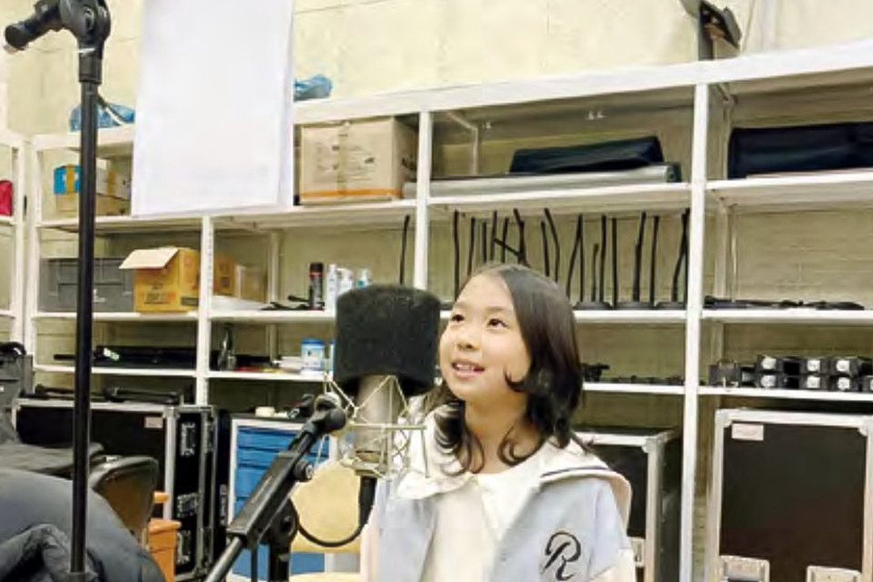 한 아이가 밝은 표정으로 마이크 앞에 서 있다. 그 뒤로 여러 장비가 놓인 선반이 있다.