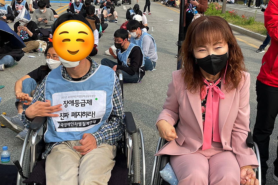 필자와 다른 한 사람이 휠체어에 앉아 있고, 뒤로 도로 한복판에 많은 사람이 앉아 있다.