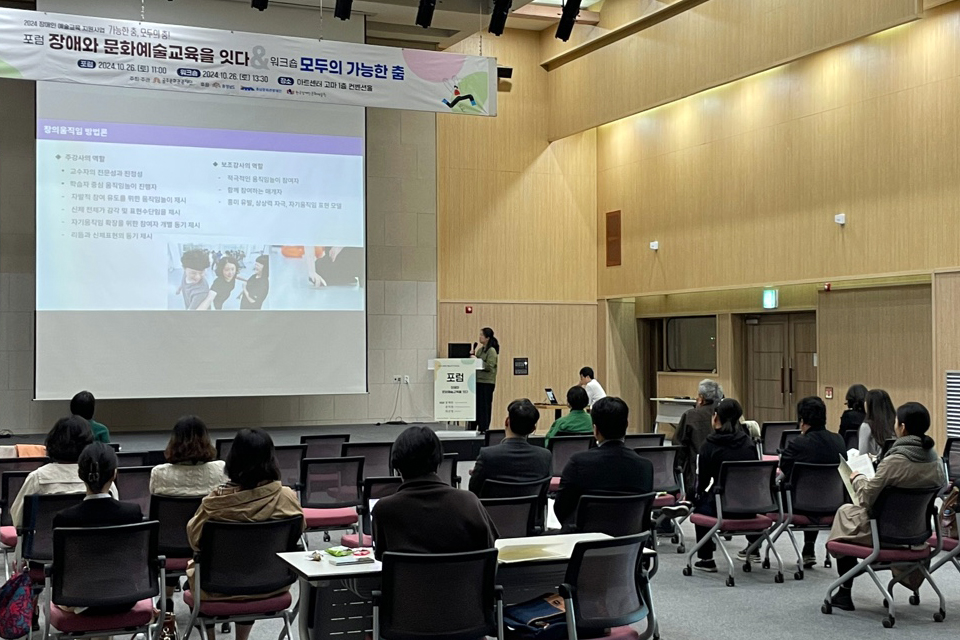 강단에서 발제자가 커다란 스크린의 내용을 소개하고 있고, 플로어에는 청중이 테이블 의자에 앉아서 듣고 있다.