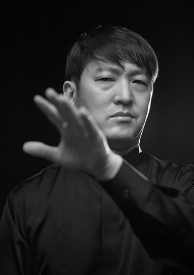 검은 배경 앞에 이훈이 서서 정면을 향해 손을 뻣으며 카메라를 응시하고 있다.