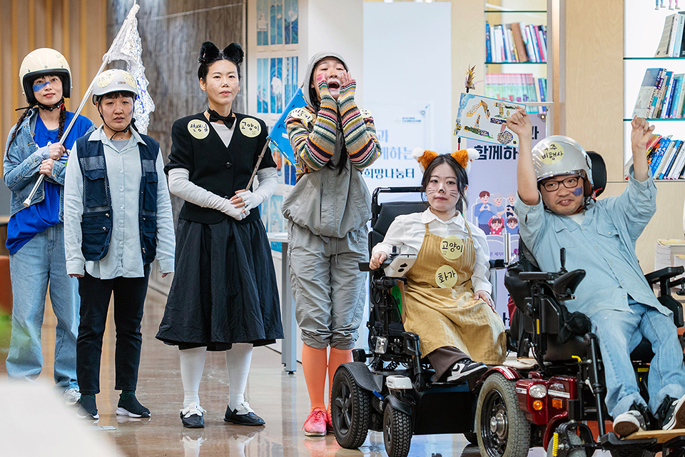 몸에 우주비행사, 고양이, 화가, 선생님, “고양이, 새벽 등의 이름표를 붙인 배우들이 휠체어를 타거나, 안전모를 쓰거나, 두 손을 모아 소리치거나, 깃발을 든 채 줄지어 앞으로 움직인다.