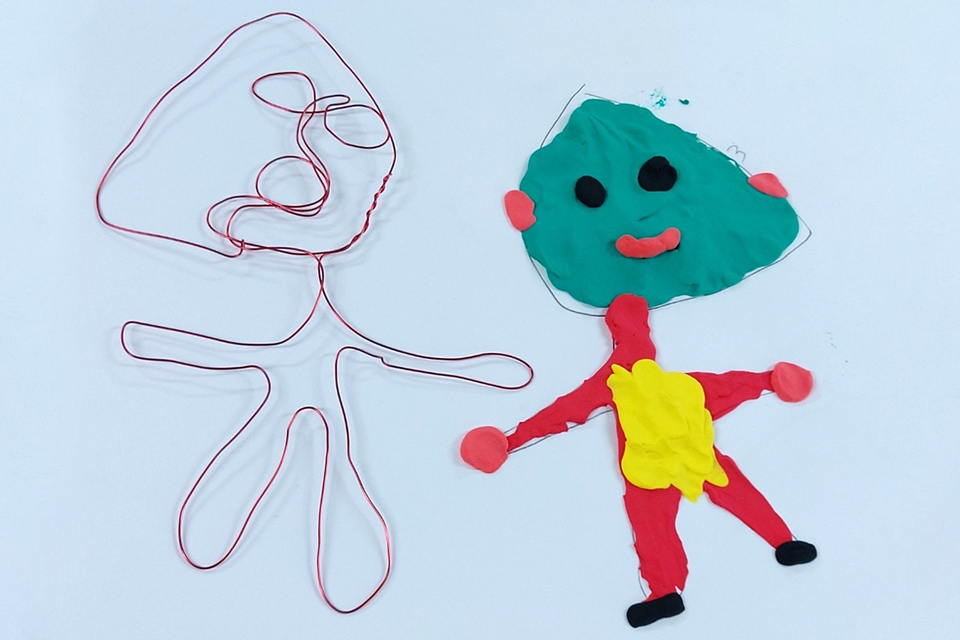 왼쪽에는 빨간 철사로 만든 추상적인 사람 형상이 있고, 오른쪽에는 찰흙으로 만든 인형이 있다. 찰흙 인형은 초록색 얼굴에 검은 눈과 입, 붉은 볼을 가지고 있으며, 빨간 팔다리에 노란색 옷을 입고 있다.