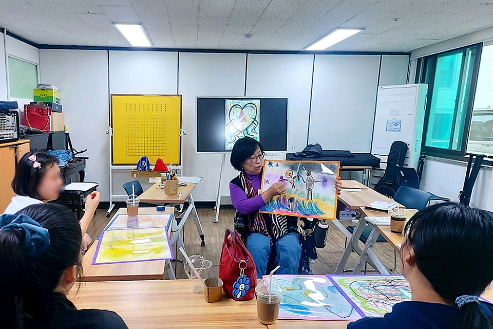 교실에서 필자가 그림을 펼쳐 보여주며 이야기를 들려주고 있고, 네 사람이 경청하고 있다. 책상 위에는 그림 자료와 음료가 놓여 있으며, 뒷편에는 칠판과 화이트보드가 세워져 있다.