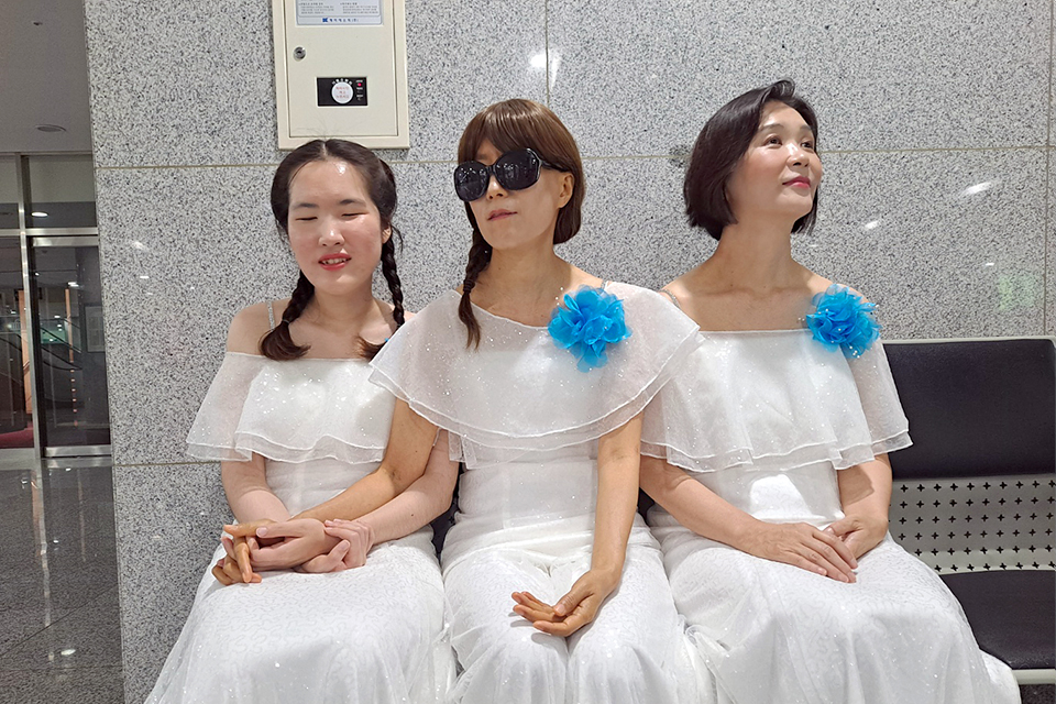 세 명의 합창단원이 흰색 드레스를 입고 건물 내부 벤치에 나란히 앉아 있다. 세 사람 모두 하얀 드레스에 파란색 꽃 장식을 달고 있다. 
