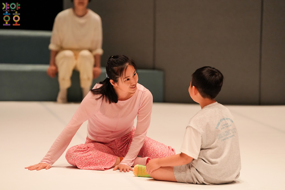 무용수가 어린이와 바닥에 마주 보고 앉아 눈을 맞추고 있다.