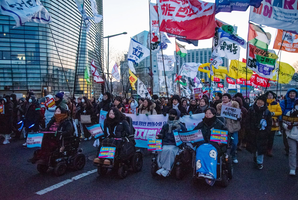 광화문 앞 도로에서 피켓을 들고 휠체어를 탄 사람들과 깃발을 든 사람들이 행진하고 있다. 