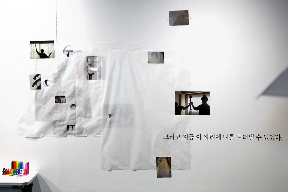 하얀 벽면에 원형과 불규칙한 모양의 구멍이 뚫린 흰 천이 붙어 있고, 구멍 사이로 여러 사진의 부분이 보인다. 사진 속에는 사람의 실루엣, 손짓 등이 담겨 있다. 사진 아래에 “그리고 지금 이 자리에 나를 드러낼 수 있었다.”라고 쓰여 있다. 하단 선반에는 오색의 레고 블록이 놓여 있다.