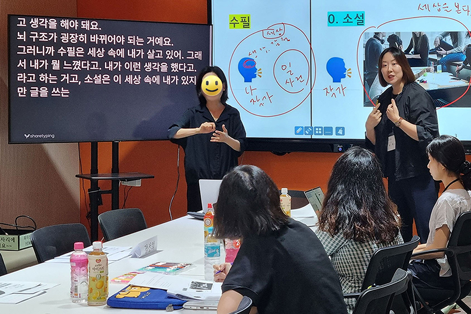 강의실에서 강사와 수어통역사가 강의하고 통역하는 중이다. 한쪽 모니터에는 자막이 띄워져 있고, 다른 모니터에는 강의 자료가 보인다. 책상에 앉은 수강생들이 이를 바라보고 있다.