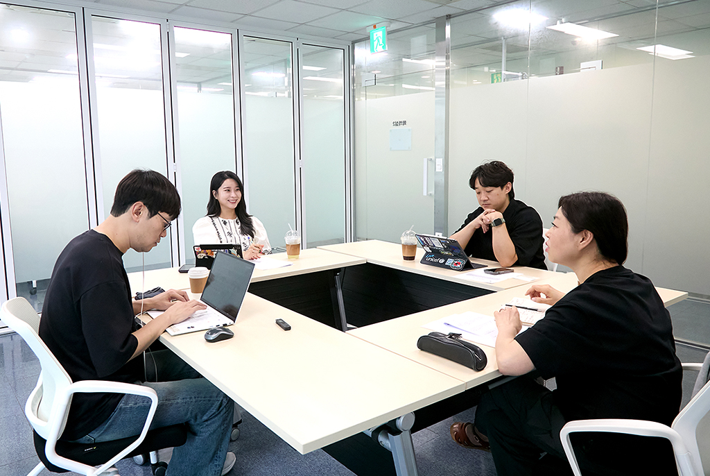 반투명 유리로 된 회의실에 테이블이 정사각형으로 배치되어 있고, 네 사람이 둘러앉아 이야기 나누고 있다. 테이블에는 노트북이나 메모지가 놓여 있다.