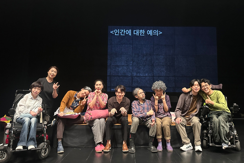 무대 중앙에 배우들이 나란히 앉아 관객을 향해 포즈를 취하고 있다. 휠체어에 탄 이도 있고, 어깨동무를 하거나 웃고 있는 모습이다. 뒷의 스크린 자막에는 〈인간에 대한 예의〉라고 쓰여 있다.