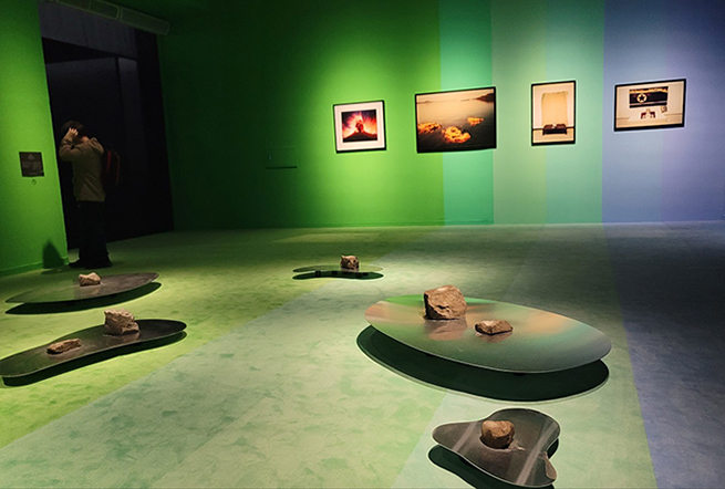 초록과 파랑이 그라데이션처럼 번지는 전시 공간. 돌을 올려둔 낮은 금속 조형물이 바닥에 여러 개 배치돼 있다. 벽면에는 자연·기억을 주제로 한 듯한 사진 작품들이 걸려 있고, 한 관람객이 작품을 바라보고 있다.