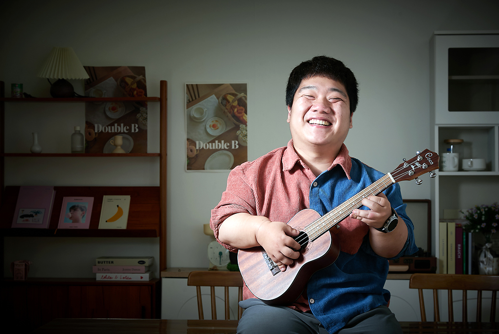 김유남 배우가 테이블 위에 걸쳐 앉아 우쿨렐레를 연주하듯 들고 활짝 웃고 있다. 반은 붉은색, 반은 파란색으로 나뉜 셔츠를 입었다. 뒤편에는 책과 소품이 놓인 선반이 있다.