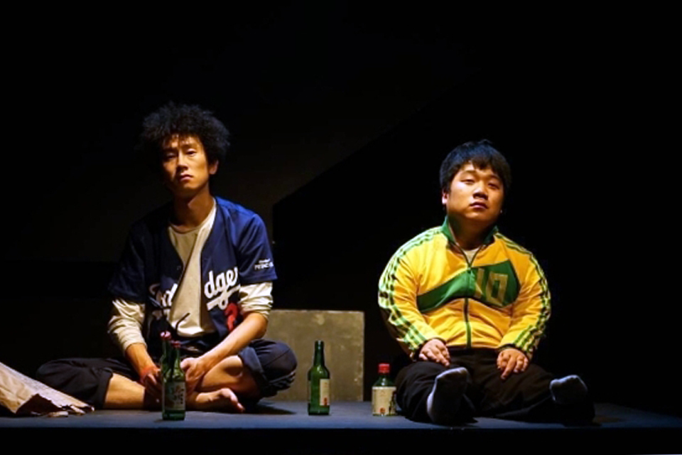 어두운 무대, 남자 배우 둘이 바닥에 나란히 앉아 허공을 바라보고 있다. 각각 파란색 야구 점퍼와 노란색 트레이닝 상의를 입었다. 앞에는 소주병과 음료수병이 놓여 있다.