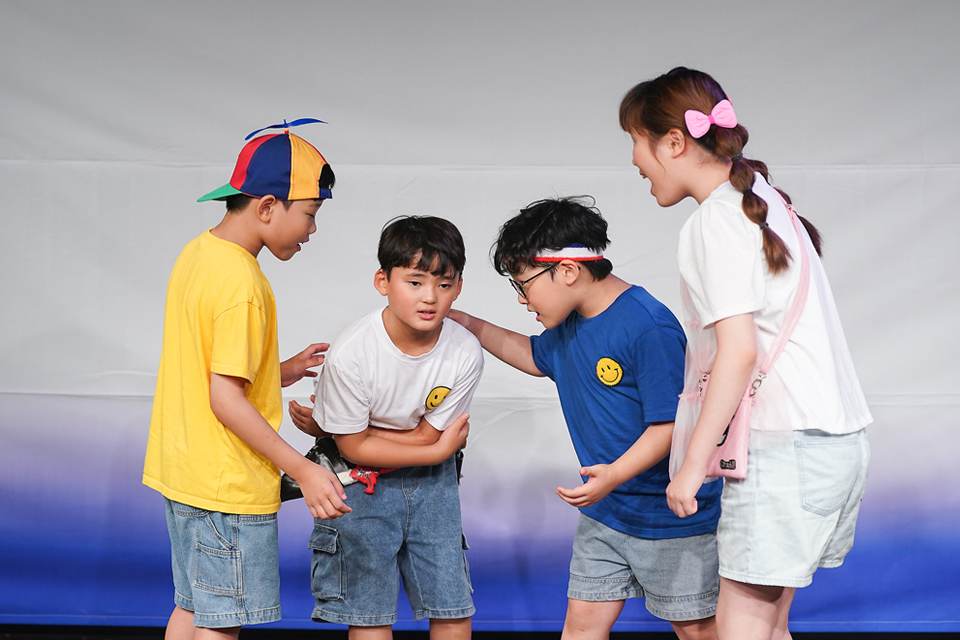 무대에 노란색, 흰색, 파란색 옷을 입은 네 명의 어린이 배우가 있다. 세 아이가 배를 감싸고 있는 한 아이를 다그치는 듯하다.