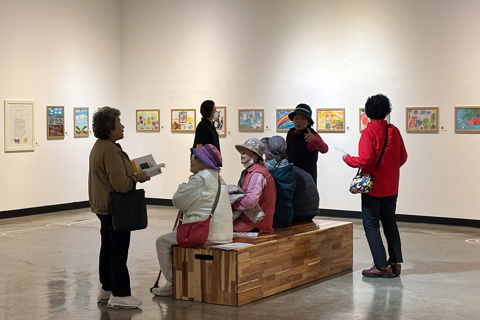 전시장 흰 벽에 여러 그림이 걸려 있고, 중·장년층의 여성 작가 7명이 벤치 주변에서 함께 전시 작품을 보거나 대화를 나누고 있다.