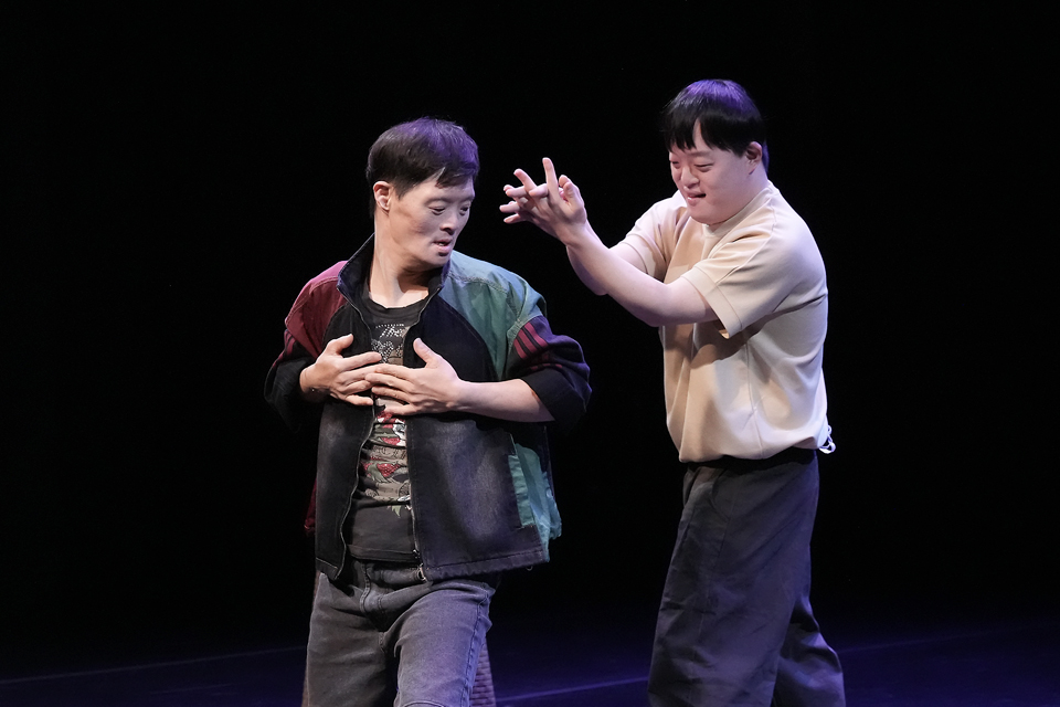 어두운 무대 위에서 김봉두가 가슴에 손을 얹고 얼굴을 김조영을 향해 돌리고, 김조영이 뒤쪽에서 두 손을 들어 함께 움직인다.