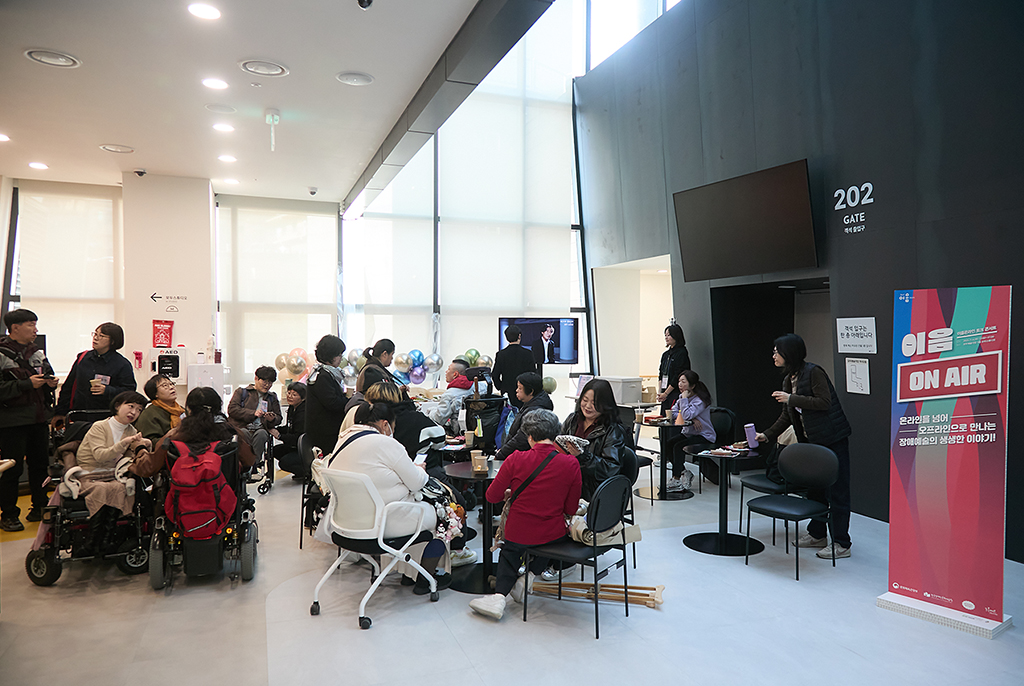밝은 로비 공간에서 많은 사람이 원형 테이블 주변에 모여 앉아 대화를 나누고 있다. 창가쪽으로 케이터링 테이블과 ‘연기가뭐라GO’가 상영되는 모니터가 놓여있다. 오른쪽에는 ‘이음온에어 ON AIR’라고 적힌 세로형 배너가 세워져 있다.