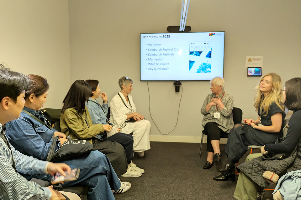 여러 사람이 벽에 설치된 모니터 좌우로 나란히 앉아 한 사람의 이야기를 듣고 있다. 모니터 화면에는 모멘텀 소개가 띄워져 있다.