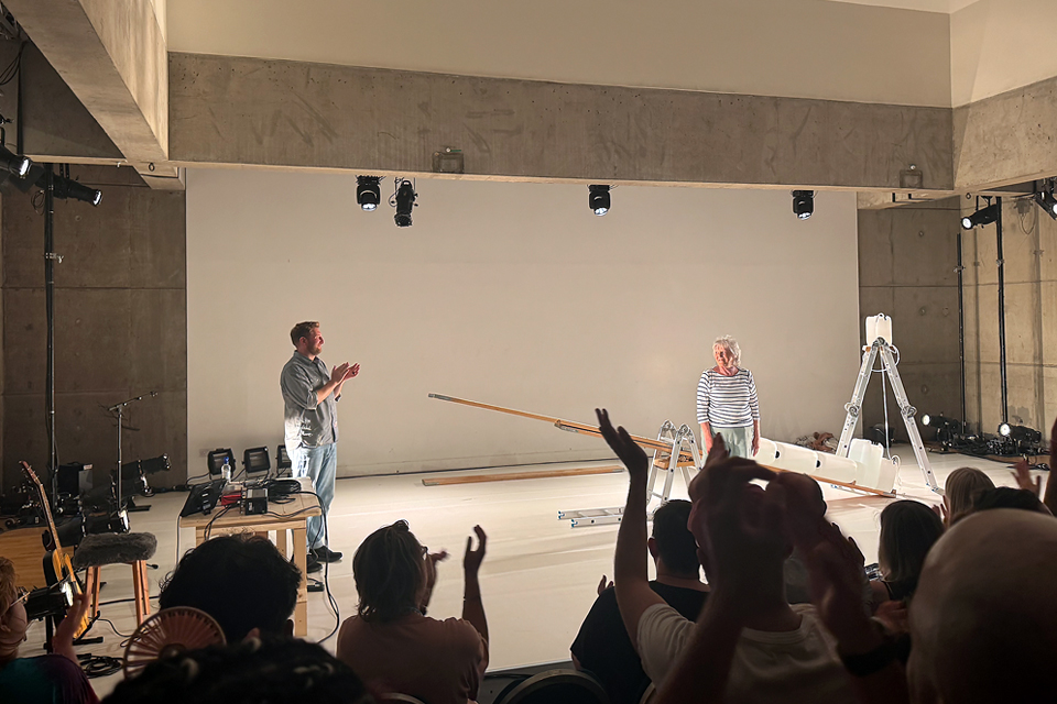 커튼콜에 두 사람이 관객을 향해 인사하고 있고, 객석에서 관객이 손을 높이 들고 박수 보낸다. 무대에는 시소, 사다리 등 기계장치들이 놓여있다.