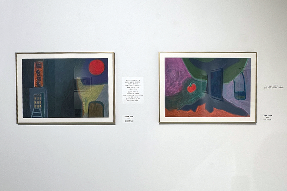 전시장 벽에 두 점의 회화 작품이 나란히 걸려 있다. 왼쪽 작품은 어두운 색면과 붉은 원형이 대비되는 구성이고, 오른쪽 작품은 보라색과 초록색이 어우러진 추상적 공간 속에 붉은 사과 모양과 병 형상이 배치되어 있다. 두 작품 사이에는 작품 설명문이 함께 부착되어 있다.