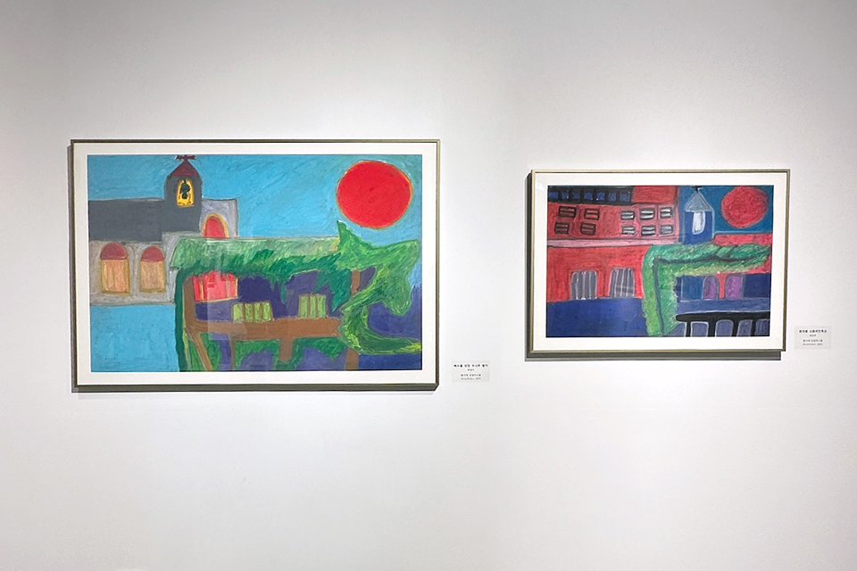 전시장 벽에 두 점의 회화 작품이 나란히 전시되어 있다. 왼쪽 작품에는 붉은 태양 아래 초록빛 풍경과 건물이 표현되어 있고, 오른쪽 작품에는 붉은색과 파란색이 대비되는 도시 풍경과 건물 형상이 그려져 있다. 두 작품은 비슷한 구도를 공유하며 색감과 분위기의 차이를 보여준다.