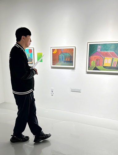 전시장 내부에서 양병철이 벽에 걸린 회화 작품들을 바라보며 천천히 걸어가고 있다. 흰 벽면에 일정한 간격으로 작품들이 전시되어 있다.