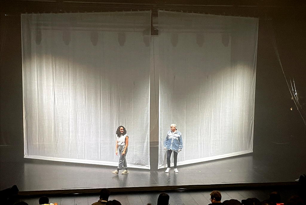무대 중앙에 반투명한 흰 천으로 둘러싸인 사각 구조물이 설치되어 있고, 두 명의 출연자가 관객을 바라보고 있다.