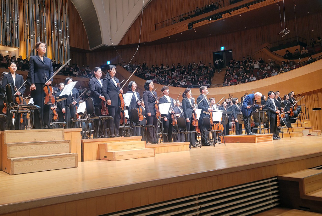 콘서트홀 무대 위에서 오케스트라 연주자들이 일제히 일어나 있고, 지휘자가 관객을 향해 인사하고 있다.