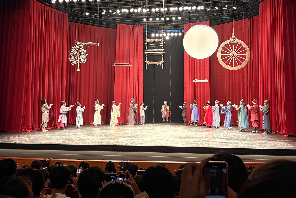 붉은 커튼을 배경으로 무대 위에 여러 명의 출연자가 일렬로 서서 공연을 마친 뒤 관객에게 인사하고 있다. 천장에는 나무·바퀴·달·의자가 매달려 있고, 객석을 가득 채운 관객들이 공연의 끝을 함께 바라보고 있다.