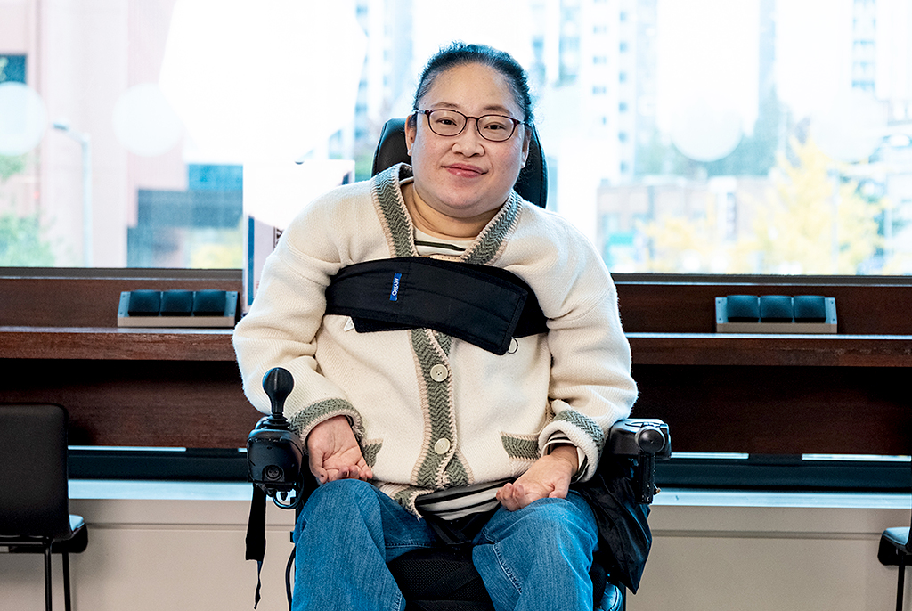 김혜정 작가가 밝은 실내 공간에서 큰 창을 배경으로 전동휠체어에 앉아 있다. 큰 창 너머로 도심 풍경과 노란 은행잎이 보인다. 작가는 안경을 착용하고 묶은 머리에 아이보리색 가디건을 입고, 얇게 미소지으며 카메라를 바라보고 있다.