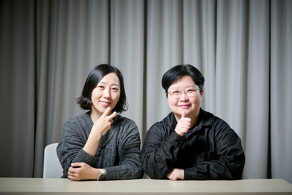 책상 앞에 조희경, 지혜원 두 사람이 나란히 앉아 카메라를 향해 미소 짓고 있다. 두 사람은 오른손을 들어 수어의 [감수], [통역] 의미를 연상시키는 동작을 하고 있다. 뒤쪽으로 부드럽게 주름이 잡힌 회색 커튼이 드리워져 있다.