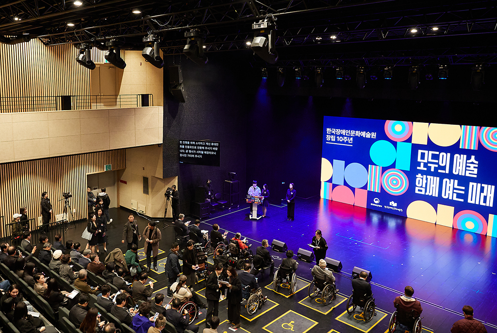 관객들이 객석을 거의 채운 가운데, 무대 오른편 대형 스크린에는 ‘모두의 예술, 함께 여는 미래’라는 문구가 밝게 표시되어 있다. 무대 왼쪽에는 진행자 두 명과 수어통역사가 있다. 주변에는 카메라 촬영팀, 자막화면, 운영 스태프 등이 있다.