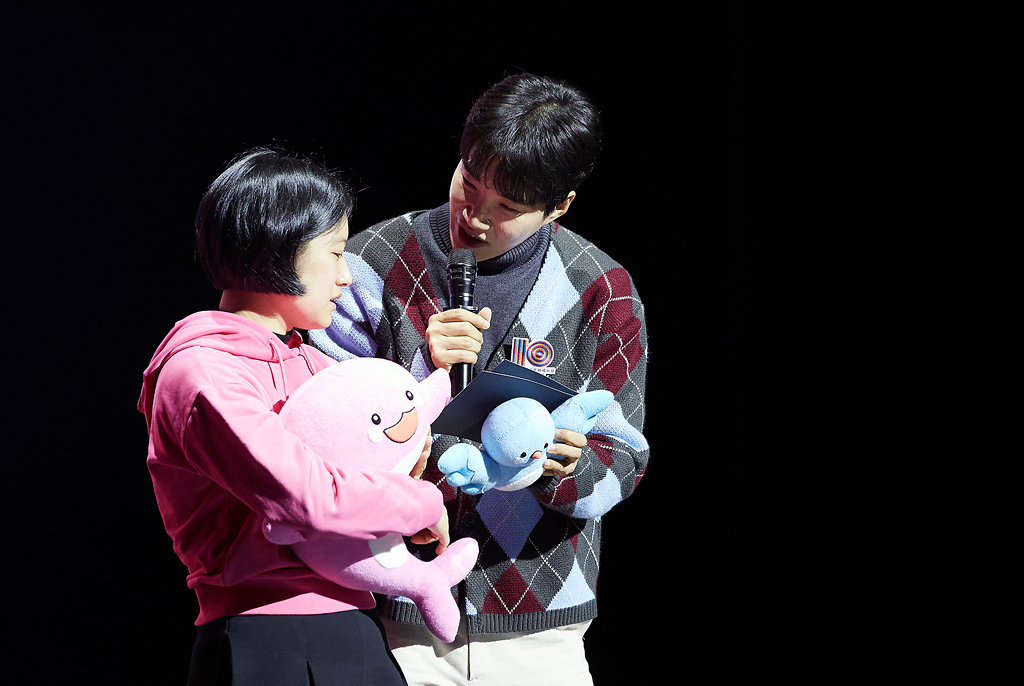 분홍색 후드티를 입은 강선아 작가는 두 손으로 고래 인형 도레를 안고 있고, 김기리 개그맨은 강선아 작가를 바라보며 한손에는 새 인형 솔라를 들고, 다른 손에 마이크를 들어 말하고 있다.