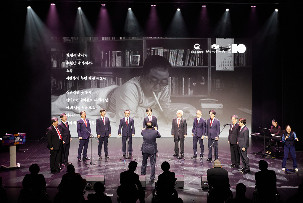 정장 차림의 단원들이 반원 형태로 서서 노래부르고, 중앙에서 지휘자가 지휘하고 있다. 뒤편 대형 스크린에는 흑백 사진으로 이상열 시인의 엎드려 입에 펜을 물고 글쓰는 모습과 시가 자막으로 보인다.