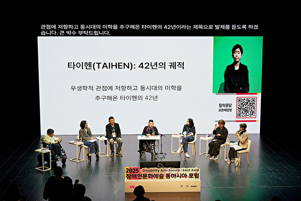 무대 위에 7명의 패널이 원형으로 앉아 있다. 뒤편 대형 스크린 상단에는 문자통역이 나오고, 가운데에 ‘타이헨(TAIHEN): 42년의 궤적-우생학적 관점에 저항하고 동시대의 미학을 추구해온 타이헨의 42년’이라는 발표 제목 텍스트가 보인다. 오른쪽에 수어통역사 영상, 질의응답을 위한 오픈채팅방 큐알코드가 띄워져 있다. 맨 앞에는 ‘2025 장애인문화예술 동아시아 포럼’ 입체간판이 놓여 있다.