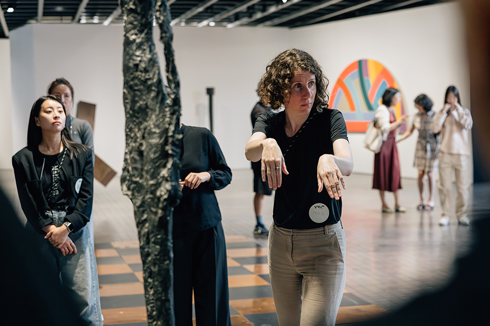 전시장에서 외국 여성 퍼포머가 팔을 굽혀 앞으로 내밀며 섬세한 동작을 선보이고 있고, 관람객들이 둘러서서 집중하고 바라보고 있다. 주변에는 조각 작품과 회화가 배치되어 있다.