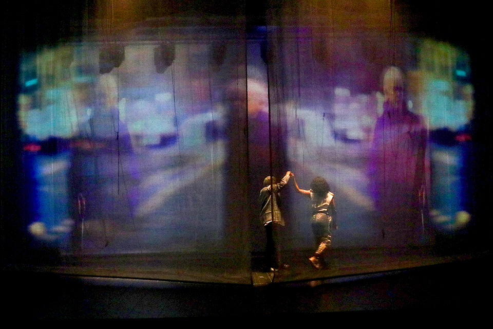 반투명한 스크린에 흐릿한 도시 풍경이 투사된 무대에서 힐다와 에바나가 서로의 손을 맞잡은 채 움직인다.